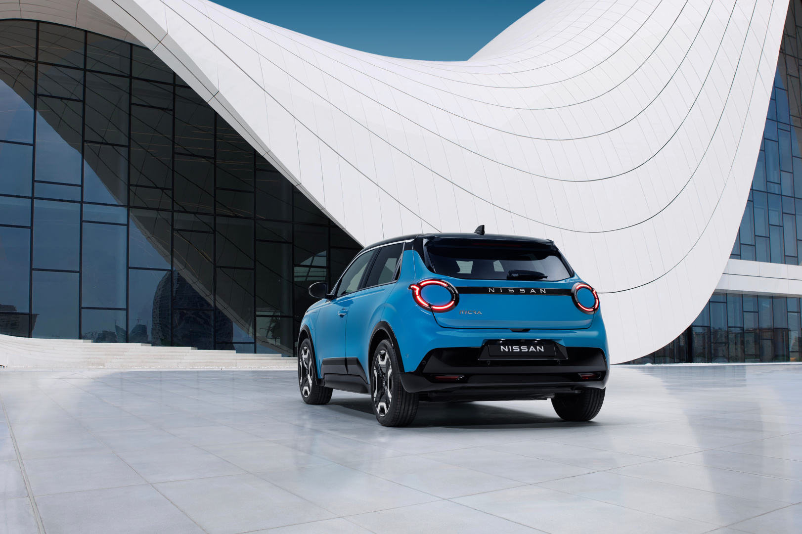 SMALL_hero shot - rear 7_8 view of new Micra in blue gear color in front of a white & blue design architectural building
